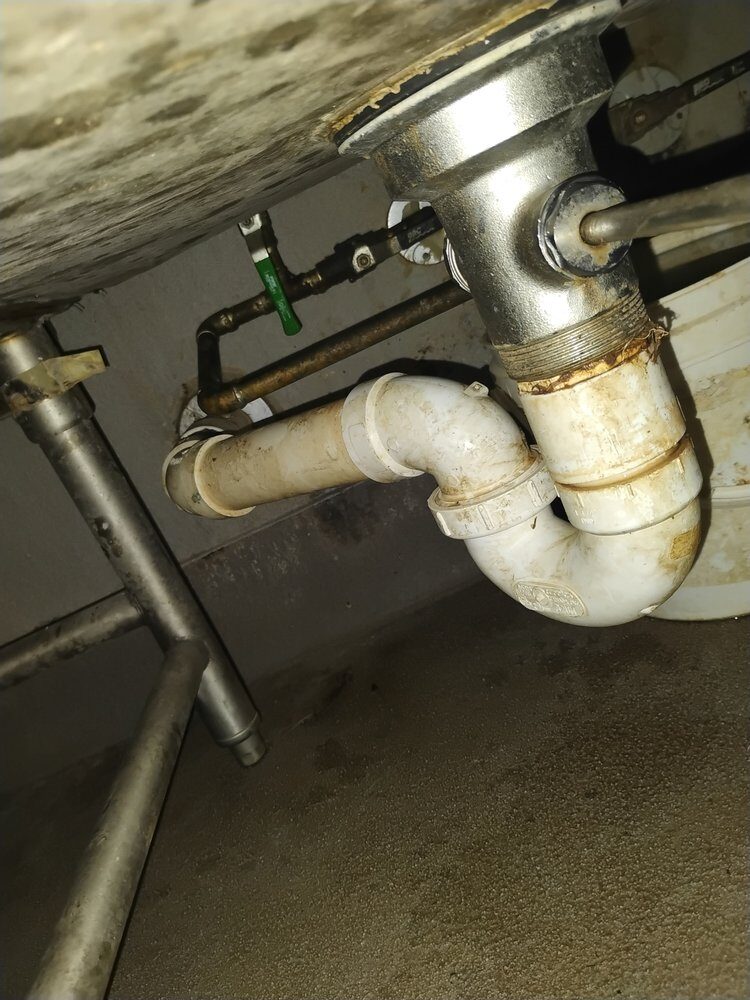 Close view of aging metal drain piping and a P-trap beneath a sink, prior to repair.
