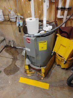 A water heater in a utility room with a damp floor and cleaning equipment nearby.